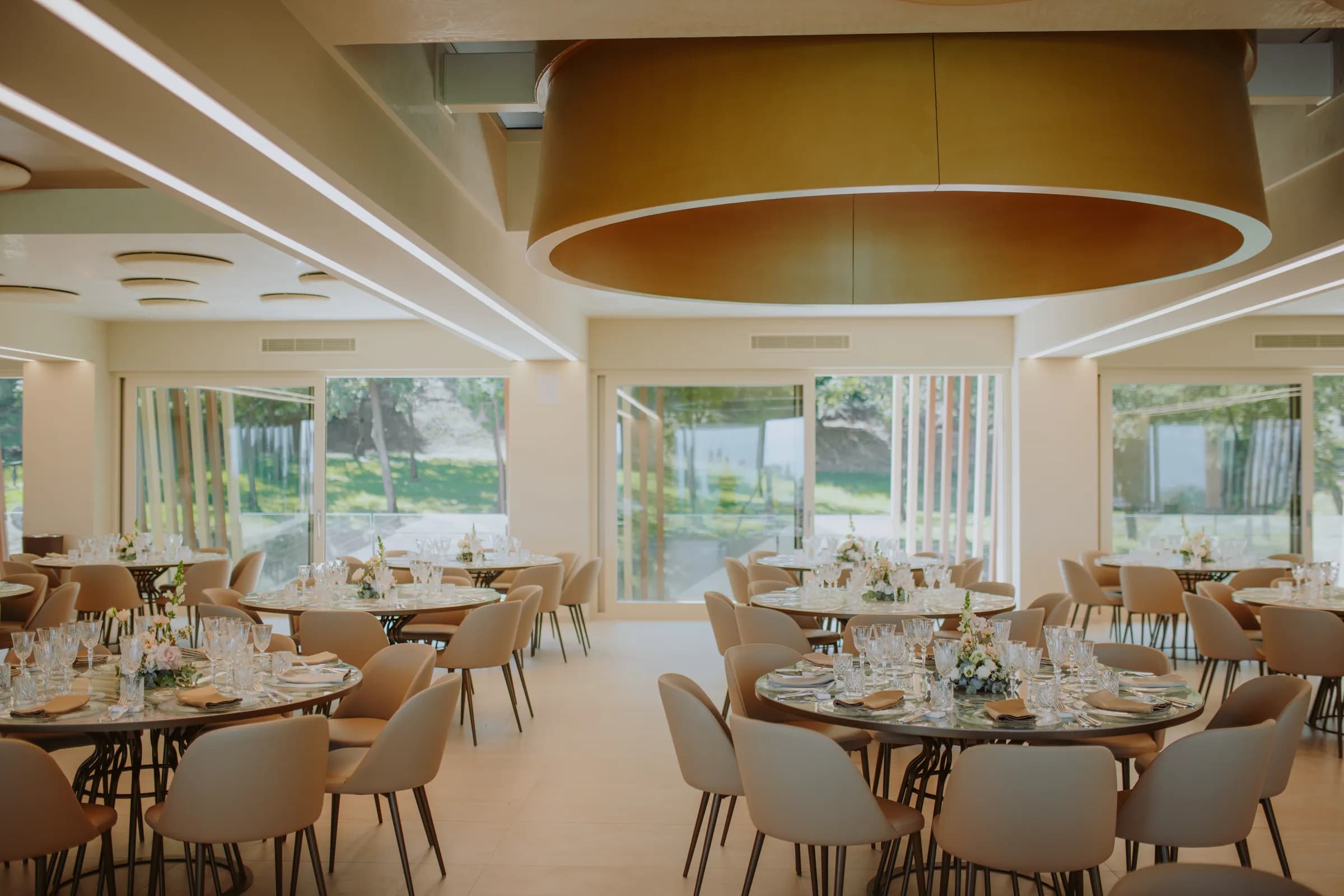 La sala apparecchiata con i tavoli tondi, sotto il grande lampadario dorato e accanto alle vetrate